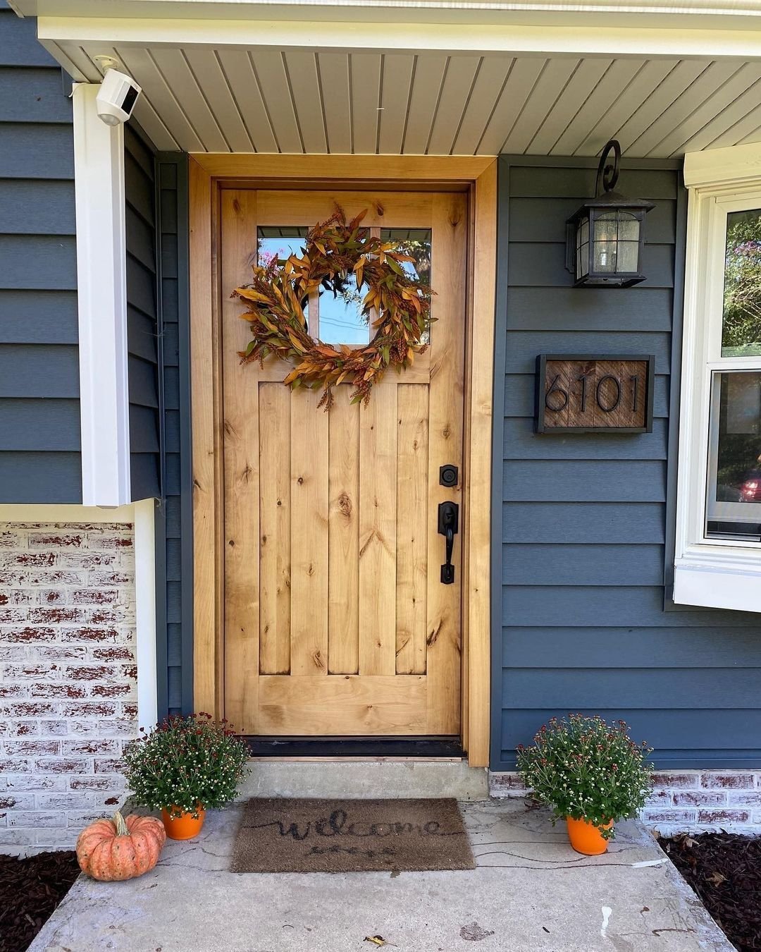 A wooden front door adorned with a fall-themed wreath, house number 8101, and a welcome mat beckons warmly. Two potted plants and a small orange pumpkin sit on either side, showcasing the versatility of solid wood front doors for every style.