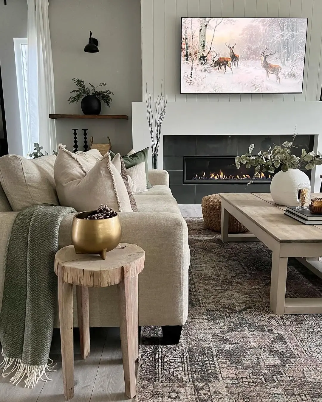 Neutral-toned living room with a beige sofa, wooden tables, and a gas fireplace creates the perfect space to embrace the season: how to transition your home decor from fall to winter. A rug, vases, throw pillows, and a TV displaying a winter deer scene complete the look.