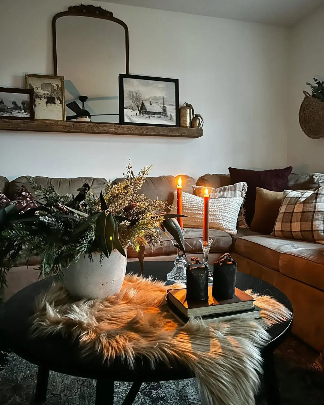 A cozy living room perfect to embrace the season: how to transition your home decor from fall to winter, featuring a beige sofa, plaid pillows, a round black coffee table with a fur throw, candles, a potted plant, and framed art on a wooden shelf.