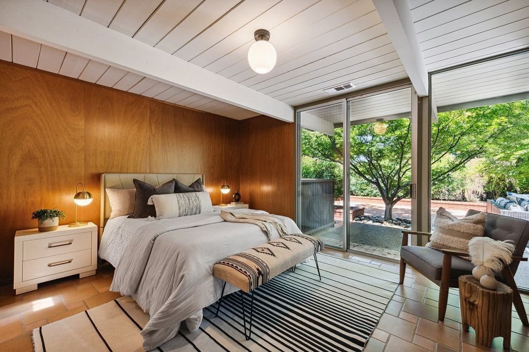 A modern bedroom with wooden walls, a large bed, and an upholstered bench epitomizes Mid Century Modern Minimalism at its best. There's a bedside table with a lamp and a small plant. Large glass doors open to a lush outdoor area with a tree, capturing the essence of Eichler Homes' seamless indoor-outdoor living.