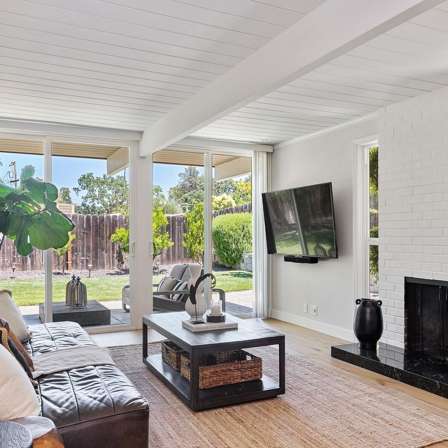 A modern living room embodies mid-century modern minimalism at its best, featuring a black leather sofa, coffee table, wall-mounted TV, and a white brick fireplace. Large windows offer a view of a backyard with greenery and outdoor seating, reminiscent of Eichler Homes' iconic design.