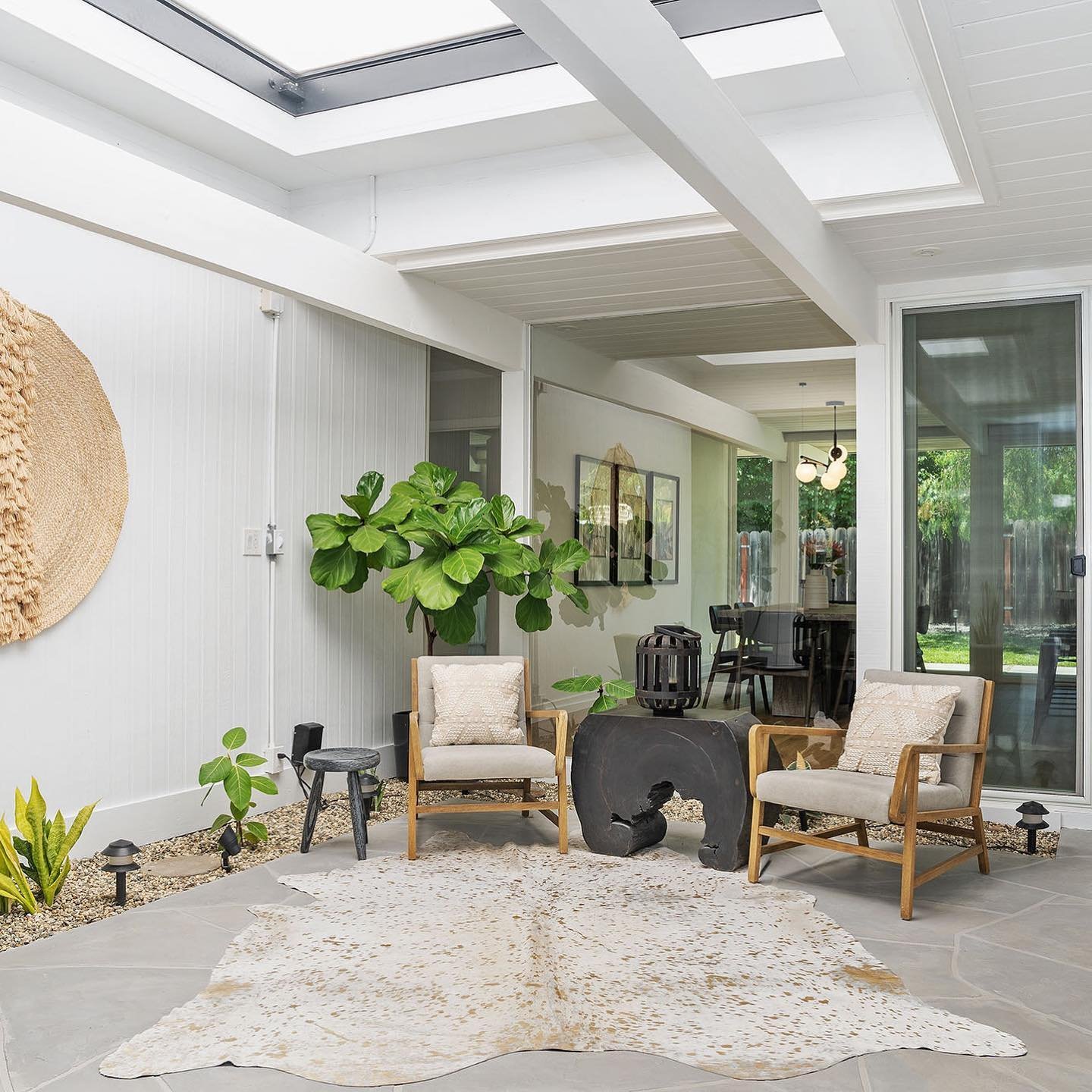 A modern sunlit room with a large skylight, two wooden chairs, a small table, a large potted plant, and a rug on the floor. A dining area is visible through a glass door in the background, showcasing Mid Century Modern Minimalism at its best: Inside Eichler Homes.