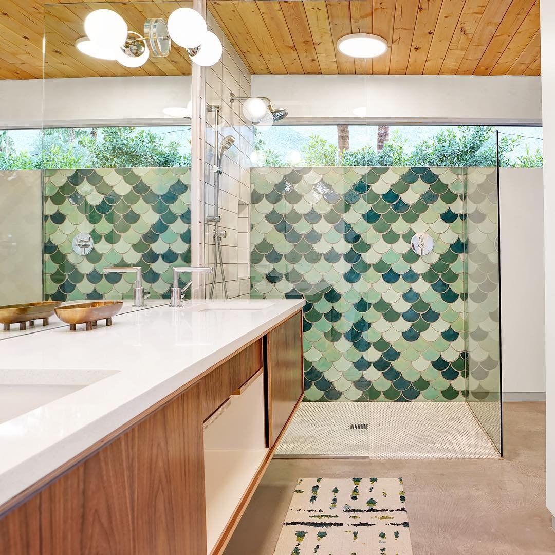 A modern bathroom features a glass-enclosed shower with green and blue fish scale tiles, a wooden ceiling, and a double sink vanity with white countertops—mid-century modern minimalism at its best.