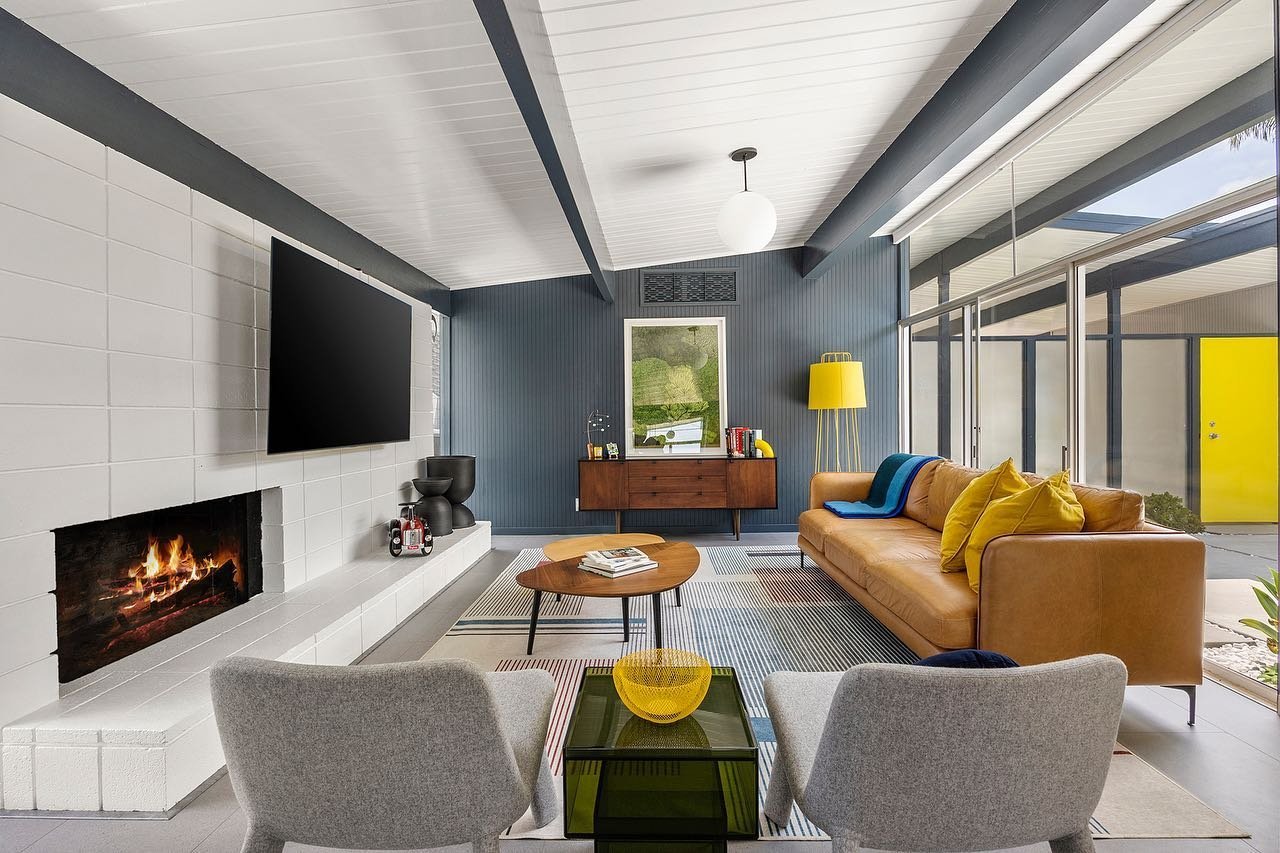 A modern living room with a fireplace, a wall-mounted TV, a leather sofa, a round wooden coffee table, two chairs, and a sideboard. Large windows bring in natural light, showcasing why Eichler Homes Interior Design is a mid-century masterpiece.