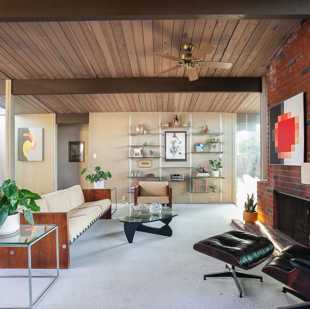 A mid-century modern living room, inspired by Eichler Homes Interior Design, features a wooden ceiling, white carpet, beige sofa, black leather lounge chair, glass coffee table, potted plants, and a brick fireplace wall adorned with framed artwork—a true mid-century masterpiece.