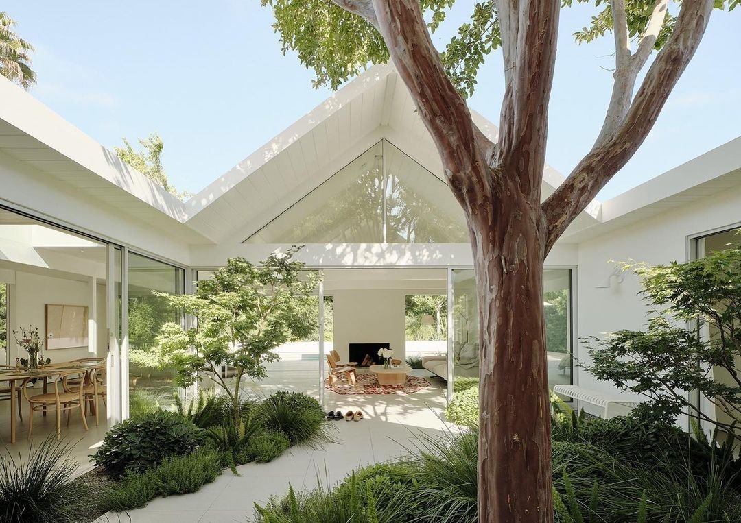 Modern home with large glass windows, landscaped courtyard with greenery and a tree in the center, and bright, minimalist interior design visible through the openings. Inspired by Eichler Homes, this design is a mid-century masterpiece that blends nature with modern elegance effortlessly.
