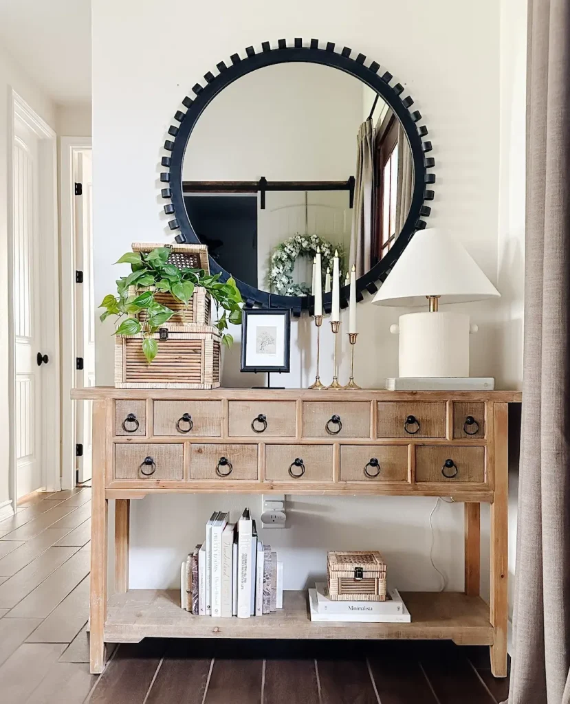 A wooden console table with multiple small drawers is adorned with plants, candles, a lamp, and books. A large round mirror hangs on the wall above the table. This setup exemplifies how a thoughtfully arranged small entryway can make a big impact using simple design hacks.