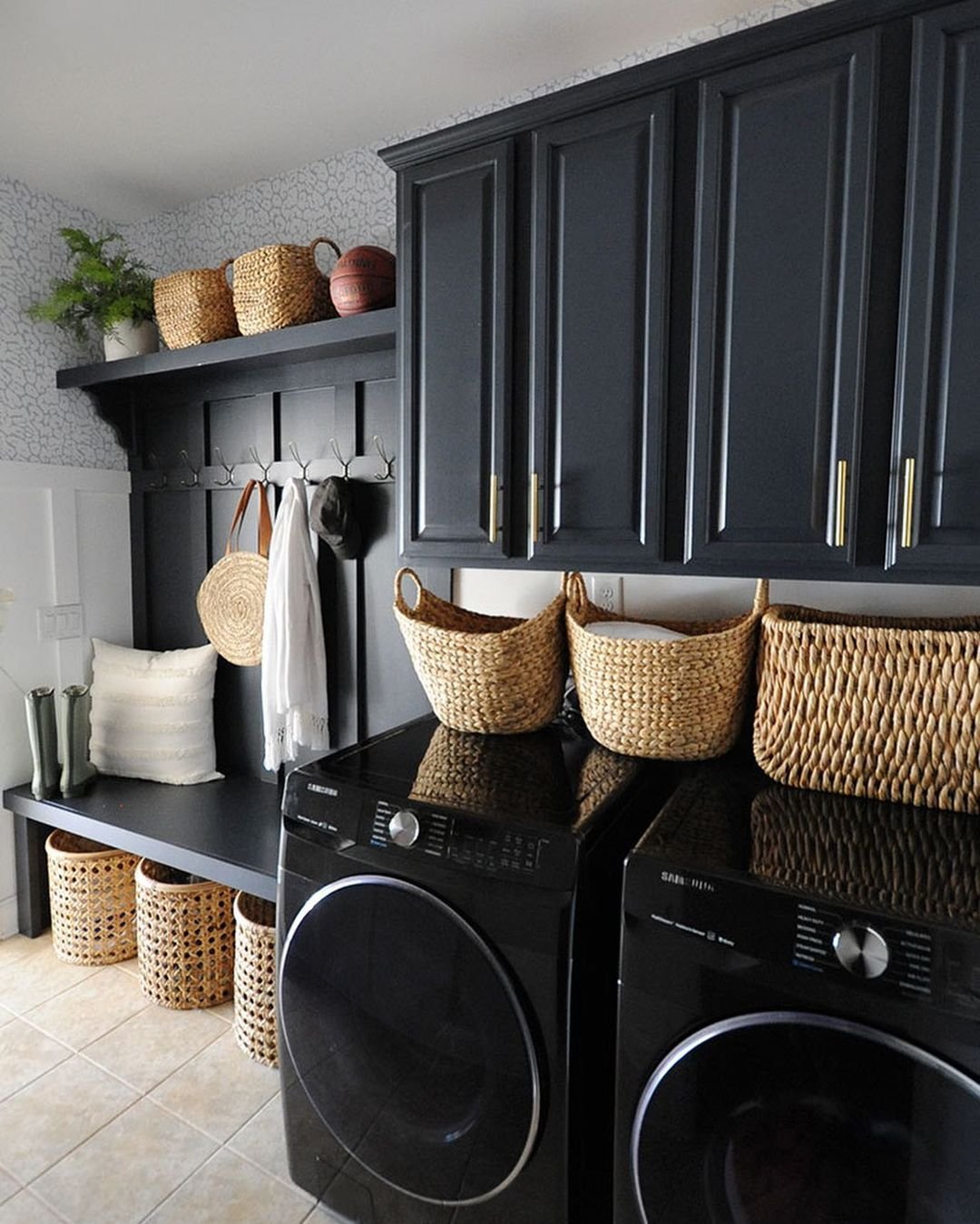 A laundry room features a black countertop over two washing machines, wicker baskets, and dark cabinets. A shelf and hooks holding hats and small items are mounted above a bench, demonstrating how creative storage hacks can transform your laundry area into an organized haven.