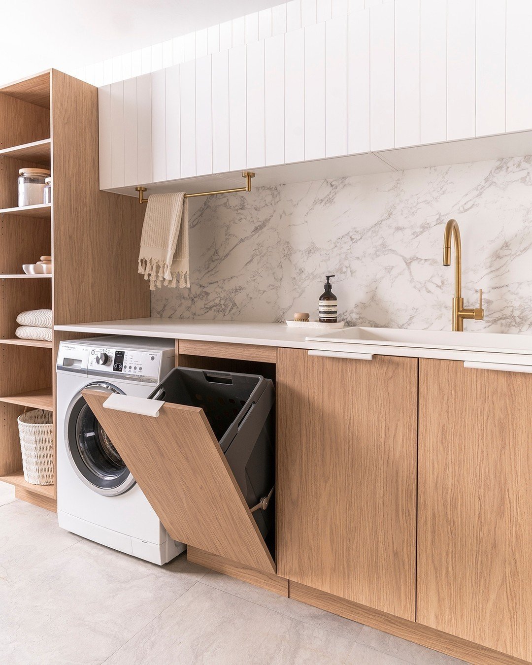 A modern laundry room with wooden cabinets, a front-loading washing machine, marble backsplash, and a pull-out bin under a white countertop with a gold faucet and hanging towel shows how creative storage hacks can transform your laundry area.