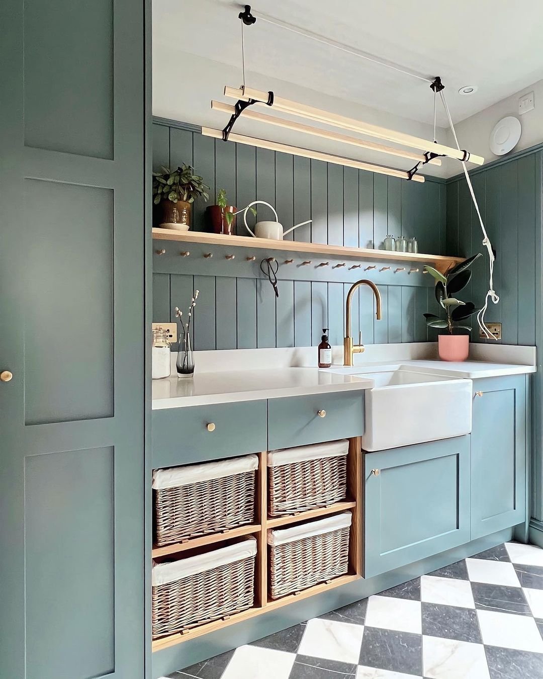 A modern kitchen with green cabinets, open wooden shelves, and a farmhouse sink showcases how creative storage hacks can elevate any space. Below the countertops are four wicker baskets in cubby holes. The floor has a black and white checkered pattern.