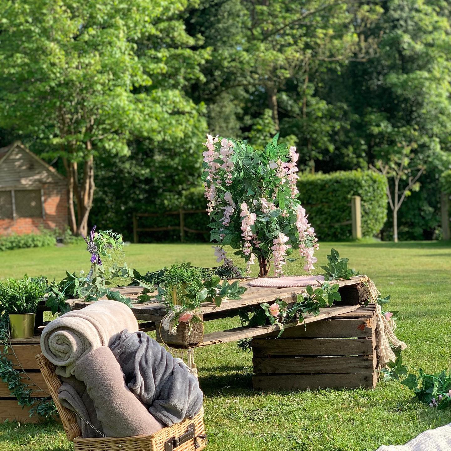 A wooden picnic table in a grassy area is adorned with pink flowers and greenery. Nearby, a woven basket with rolled blankets and a towel sits by 10 wooden crates, offering decor hacks to add rustic charm to your home. Trees and a shed create a serene backdrop.