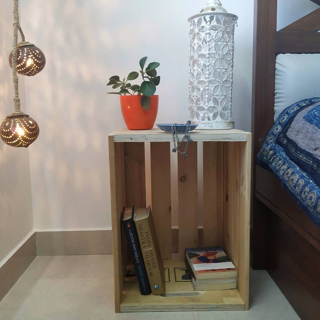 A simple nightstand made of a wooden crate holds a white decorative lantern, a small plant in an orange pot, and books on the bottom shelf. A bed with a blue and white bedspread is next to it, embodying one of the 10 Wooden Crates Decor Hacks to Add Rustic Charm to Your Home.