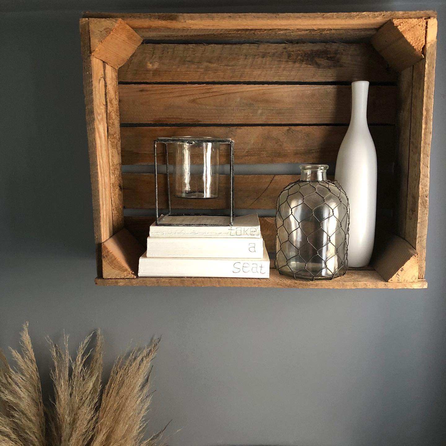 Wooden box shelf with decorative items including white and silver vases, a glass candle holder, and three stacked books against a grey wall. Pampas grass is visible at the bottom left corner, showcasing one of the 10 Wooden Crates Decor Hacks to Add Rustic Charm to Your Home.