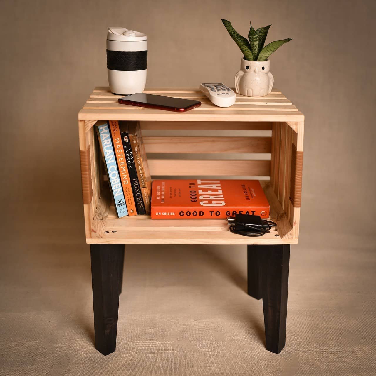 A wooden side table with black legs holds a potted plant, a remote control, a reusable coffee cup, and a smartphone on top. Books and headphones are stored on the shelf below, adding rustic charm reminiscent of wooden crate decor hacks.