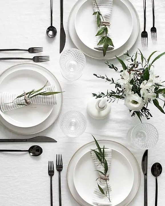 A neatly arranged table setting captures Minimalist Magic: white plates, striped napkins tied with twine and greenery, black cutlery, clear glasses, and a white floral centerpiece create simplified winter table decor on a crisp white tablecloth.