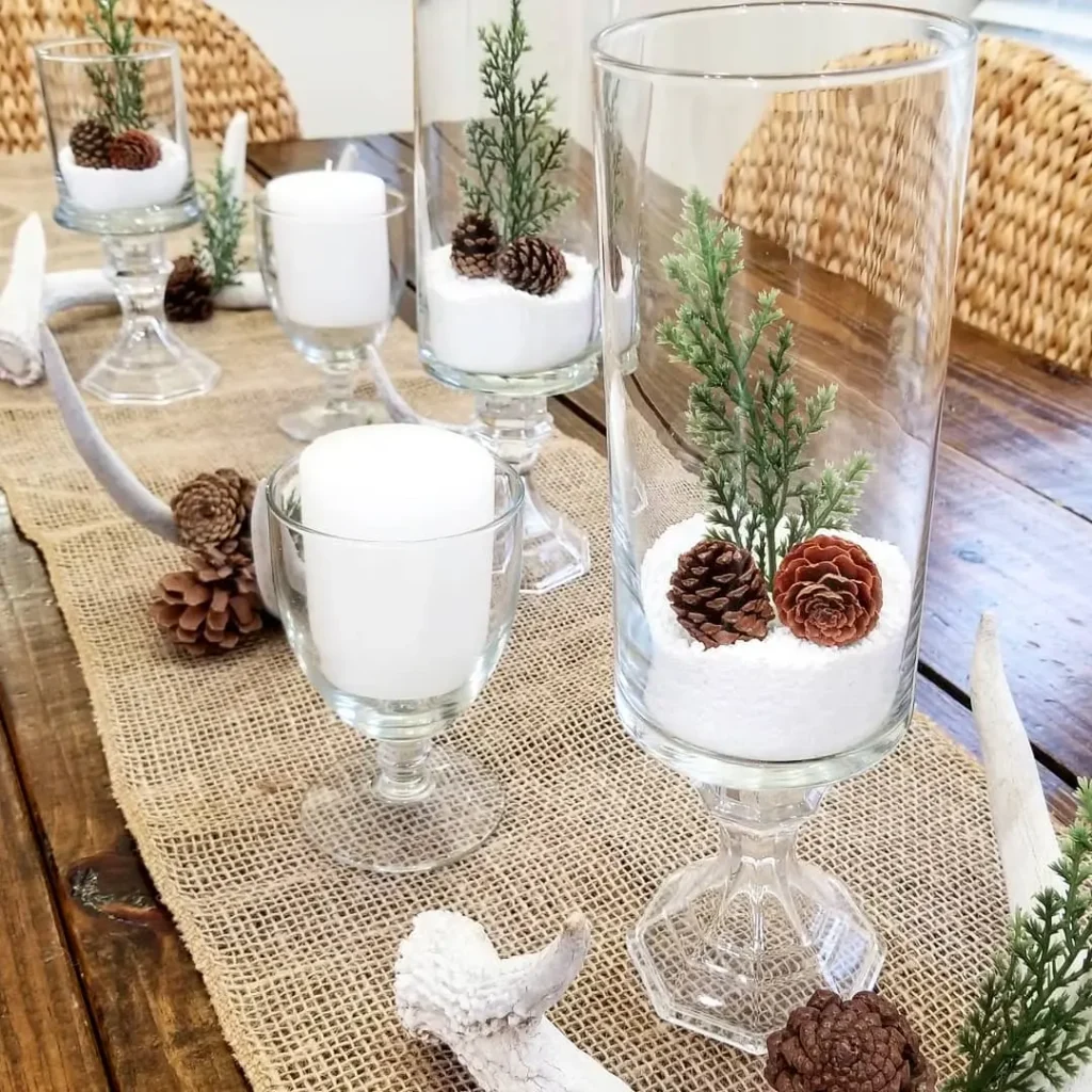 Minimalist Magic: How to Style a Simplified Winter Table Decor with glass candle holders, white candles, pinecones, artificial greenery, and faux snow arranged on a burlap runner with antler and pinecone accents.