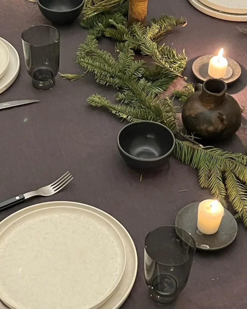 A table set with white plates, black bowls, clear glasses, candles, and pine branches on a dark tablecloth captures the essence of Minimalist Magic: How to Style a Simplified Winter Table Decor.