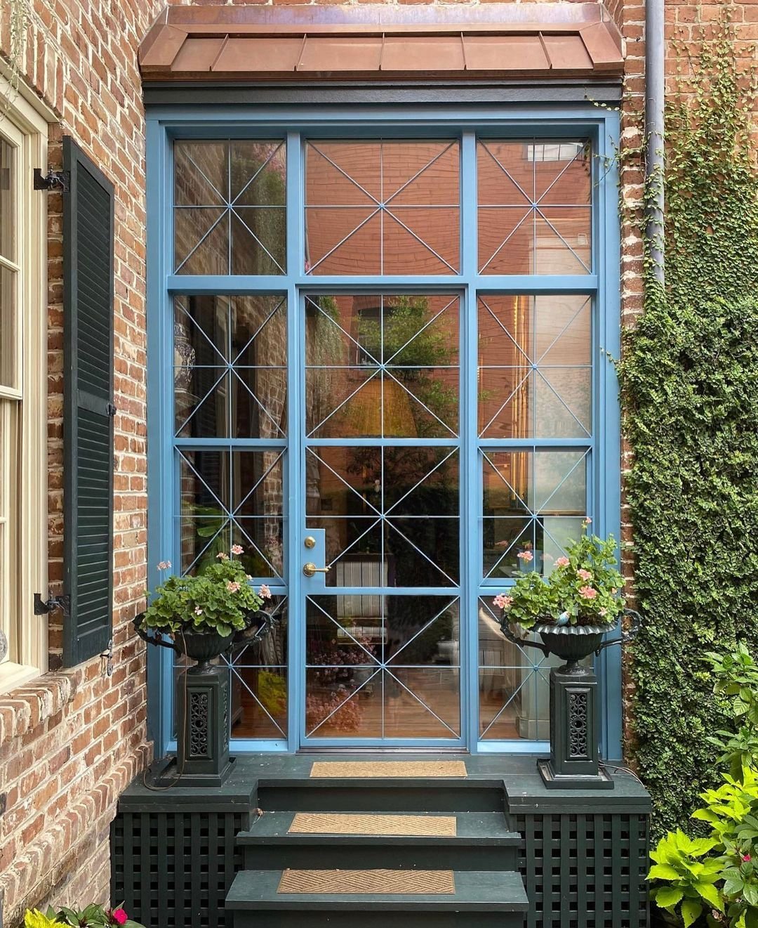 A blue-framed glass door with grid patterns is flanked by brick walls and greenery, reminiscent of steel front doors found in industrial style guides, leading to a small entryway with potted plants on either side.