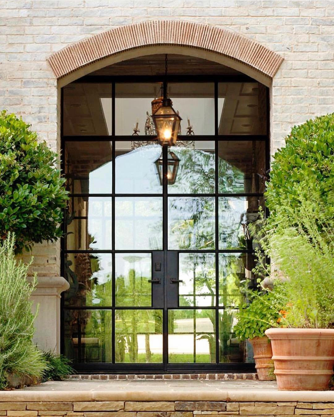 Glass-paneled steel front door with an arched, brick-framed entrance, potted plants, and a central lantern hanging from the ceiling offers an industrial style guide to chic home design.