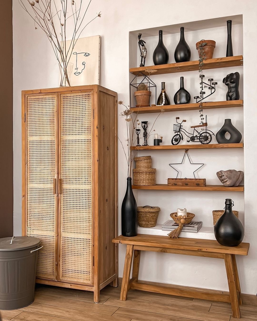 A wooden cabinet stands next to a wall with three shelves displaying black vases, small sculptures, and potted plants—an inspiring example of how to avoid common mistakes when decorating small spaces. Below the shelves sits a wooden bench.