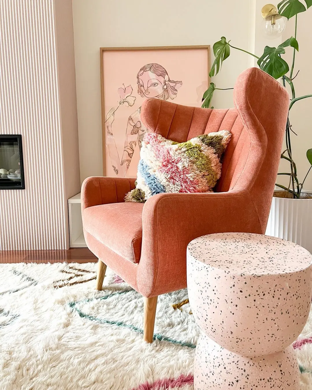A pink armchair with a colorful cushion sits next to a speckled side table on a white rug—funky furniture like this adds unique character to your space. A framed artwork and a potted plant complete the standout look.