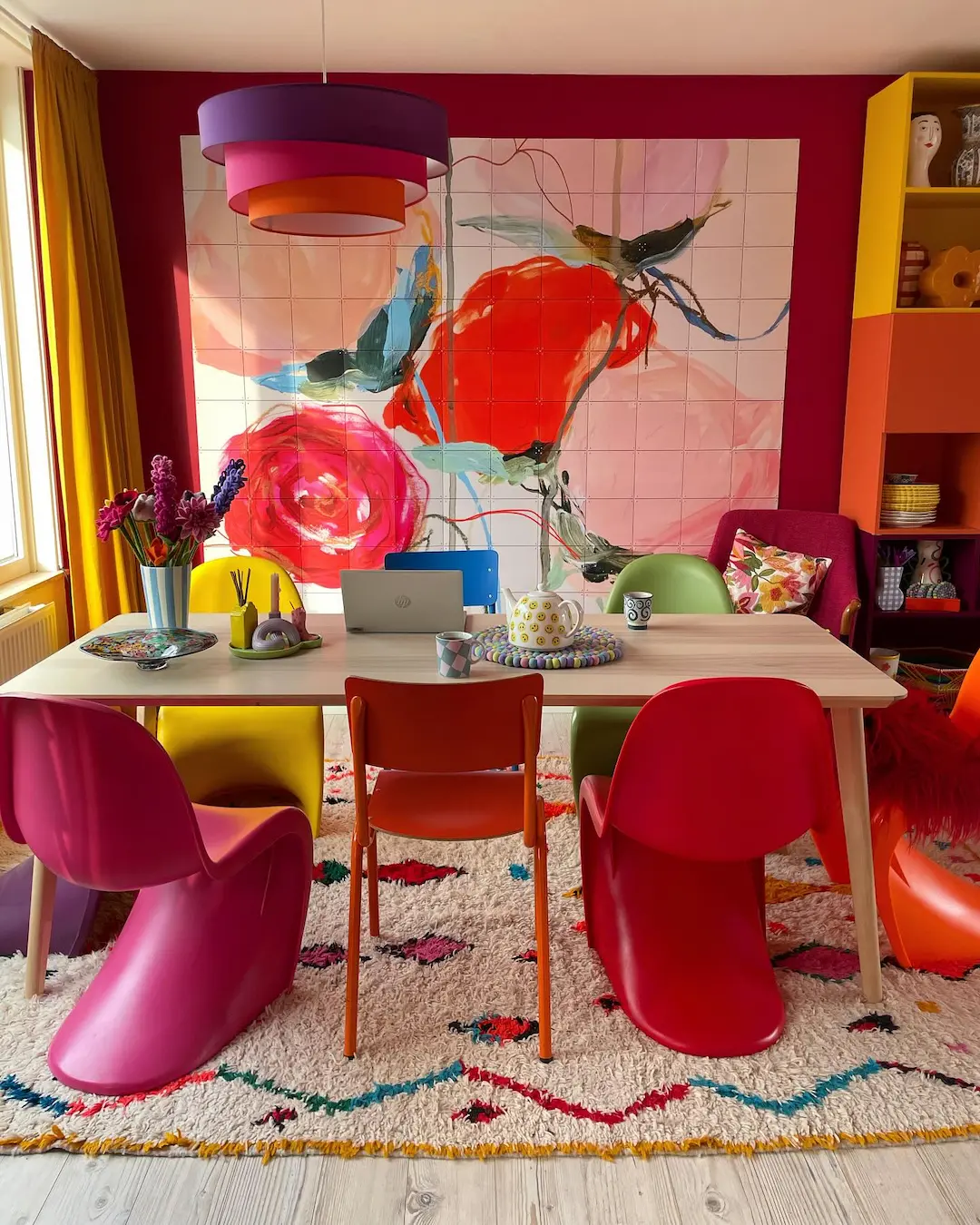 Colorful dining room with red, pink, and green chairs, a wooden table with a laptop and mugs, funky furniture accents, a vibrant abstract wall mural, and multicolored decor—unique pieces to make your home stand out.