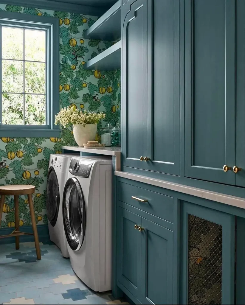 A laundry room with blue cabinets, a window, a washer and dryer, a wooden stool, and bright floral wallpaper shows how to create a moody laundry room effortlessly.