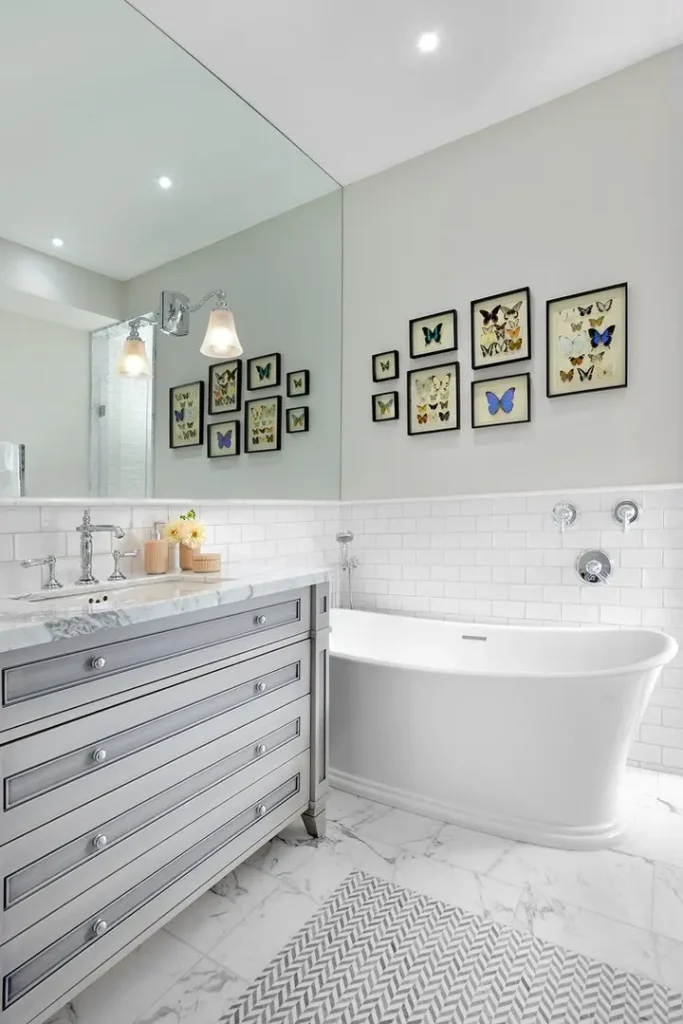Modern bathroom with a large mirror, framed butterfly artwork creating a stunning gallery wall, a freestanding bathtub, white subway tiles, marble floor, and a silver-colored vanity with a striped rug in front.