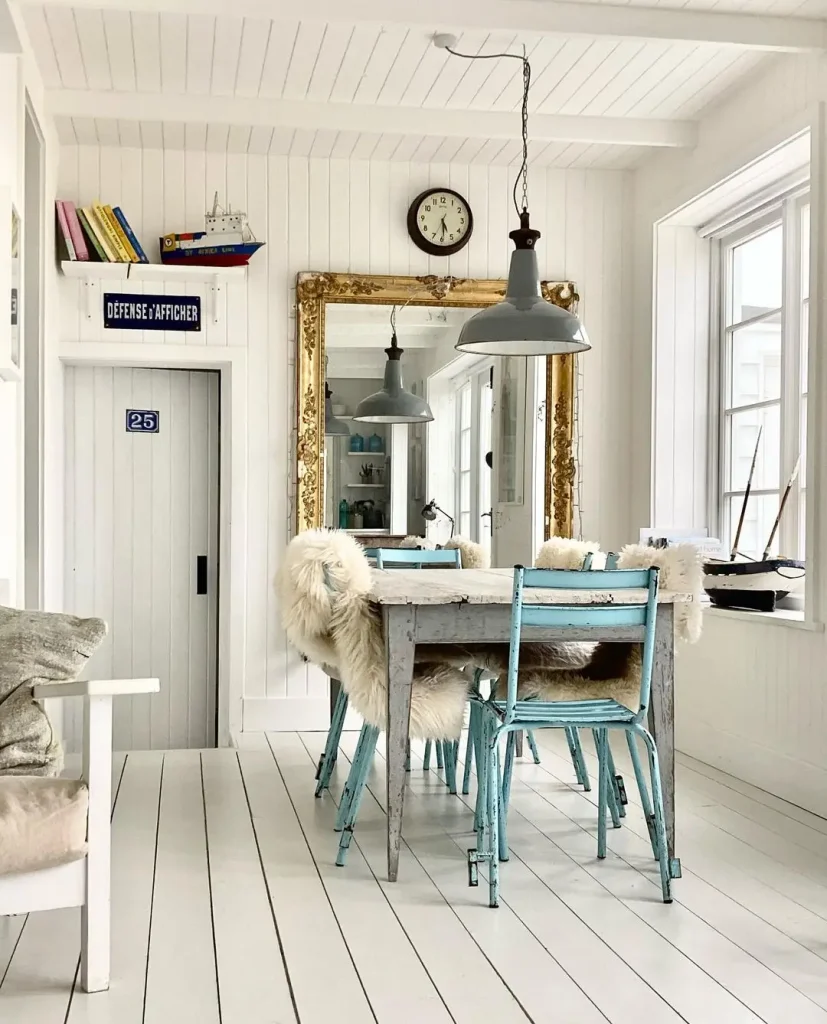 Bright dining room with white walls and floors, a rustic wooden table with blue chairs, and a large gold-framed mirror. The space showcases elements from Beach Decor 101, Everything You Need to Know to Get Started, featuring hanging gray lamps, a clock on the wall, and a door marked "25" on the left.