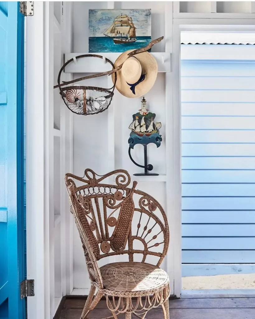 A wicker chair sits in a bright room with a hat, seashell basket, and ship model on the wall behind it. A painting of a sailing ship hangs above. This setting illustrates how to design a beach-styled living room on a budget, with white and blue walls adding to the airy coastal vibe.