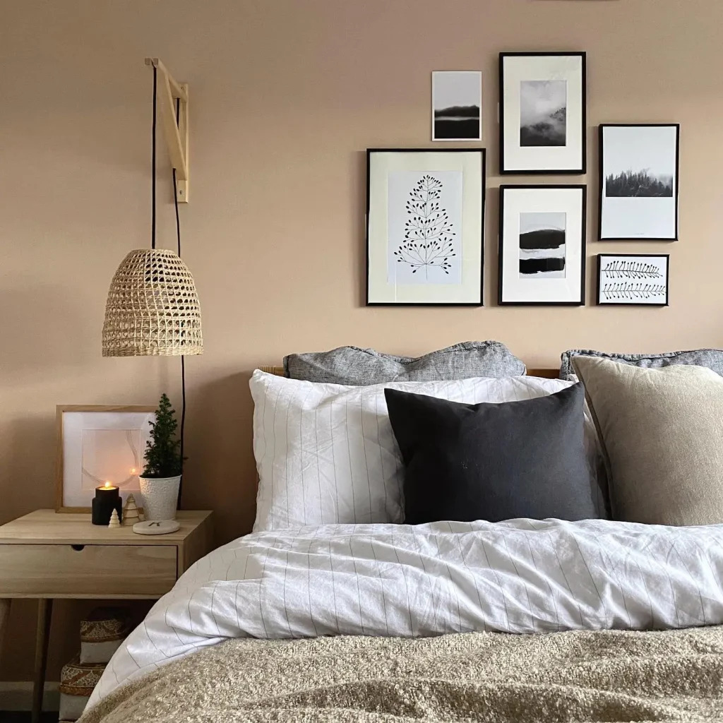 A neatly made bed with white, gray, and beige pillows and blankets sits below a stunning gallery wall. A small wooden nightstand holds a plant, a candle, and a book. Several framed pictures hang above the headboard, showing how to create a stunning gallery wall for every room.