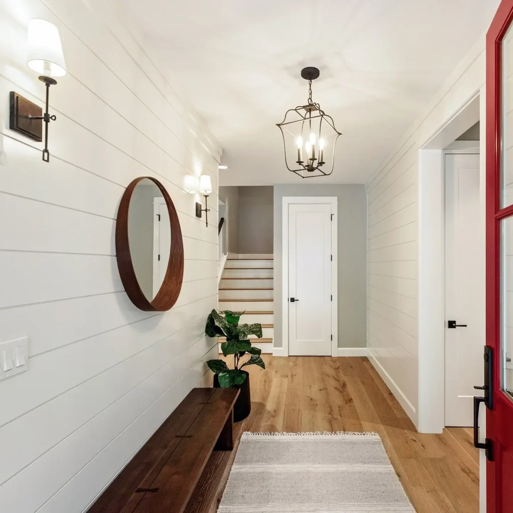 A bright, clean hallway with shiplap walls features a round mirror, wall sconces, a wooden bench, a plant, and a rug. Perfect for maximizing space in your small entryway with big impact design hacks! A staircase with a white door is at the far end, under a chandelier.