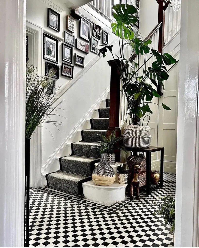 A hallway featuring a staircase with carpeted steps, framed art on the wall showcasing ideas on how to create a stunning gallery wall for every room, and various potted plants. The floor has a black and white checkered pattern.