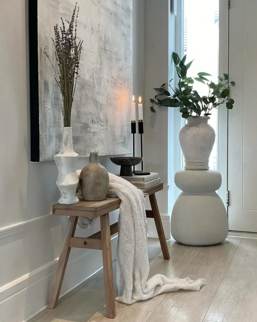 A wooden bench with a white blanket draped over it, displaying ceramic vases and a sculpture, stands near a large abstract painting. Two lit candles and a plant in a white pot add ambiance to this stylish small entryway with big impact.