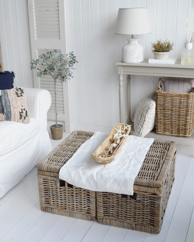 A living room with a white armchair, wicker coffee table, tray, console table, lamp, basket, and potted plant against white paneled walls shows how to design a beach-styled living room on a budget.