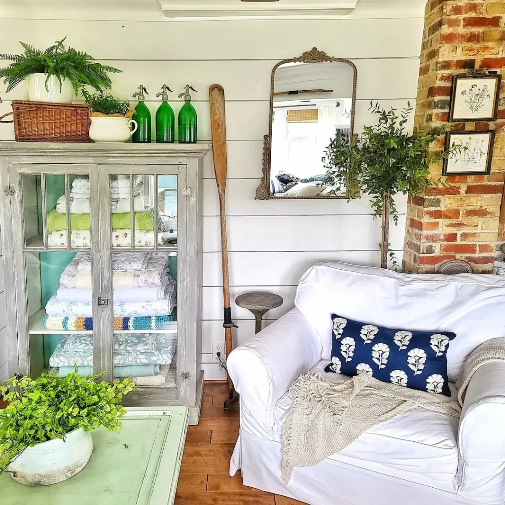A cozy room with beach decor featuring a white armchair, blue cushion, green plant on a coffee table, cabinet with folded linens, green bottles on top, a mirror, and a brick accent wall—showing how to select colors and materials for your home.