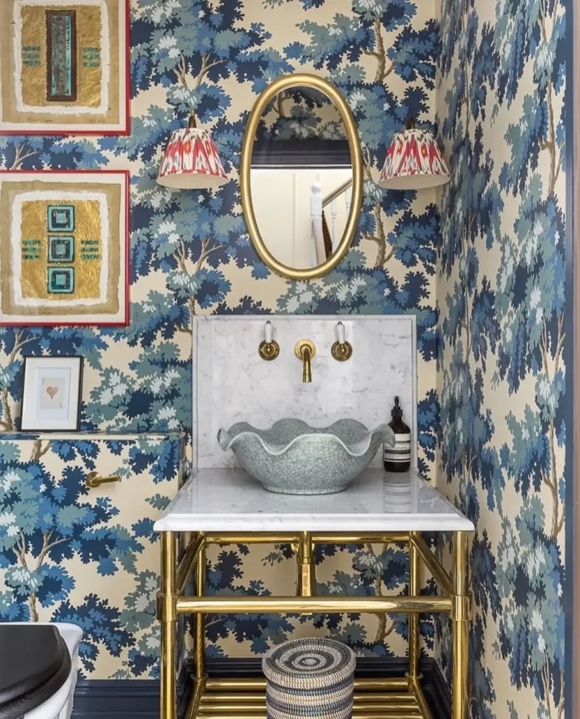 A small bathroom with blue patterned wallpaper, a marble sink vanity with gold fixtures, an oval mirror, and two wall lamps with red and white shades—perfect inspiration for Maximalist Decor: Your Ultimate Guide to Bold Living Spaces.