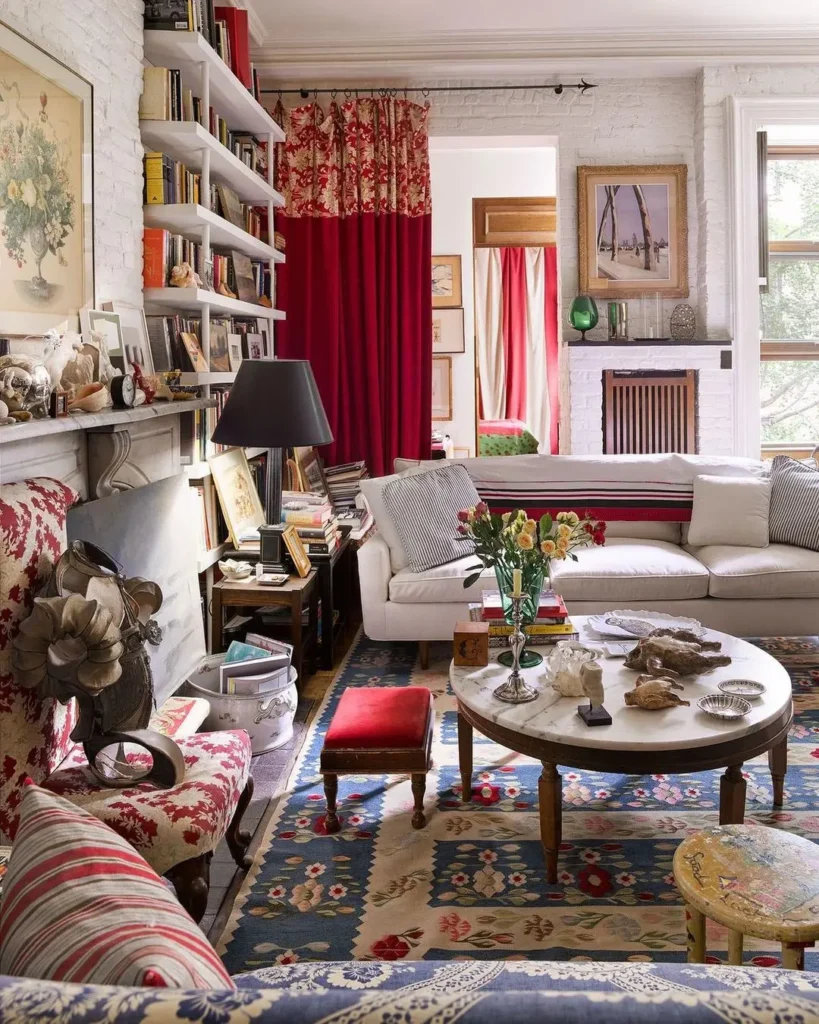 Maximalist decor brings this cozy living room to life, featuring a patterned rug, white sofa, vibrant red curtains, and eclectic decorative items on shelves and tables. A fireplace adorned with artwork and a large window that floods the space with natural light complete the bold aesthetic.