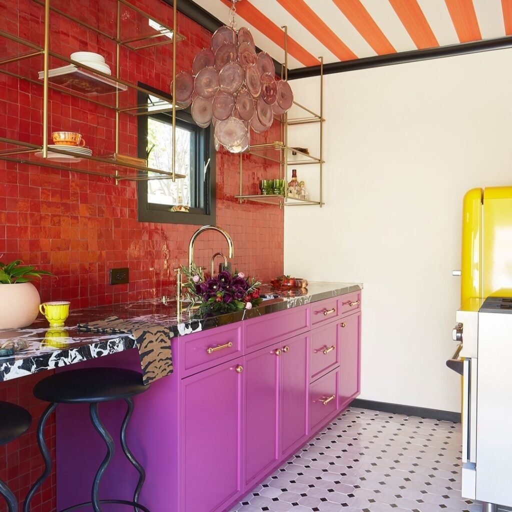 A colorful kitchen inspired by Maximalist Decor: Your Ultimate Guide to Bold Living Spaces features pink cabinets, a yellow refrigerator, red tiled walls, black and white floor tiles, open brass shelves, and a chandelier with round glass accents.