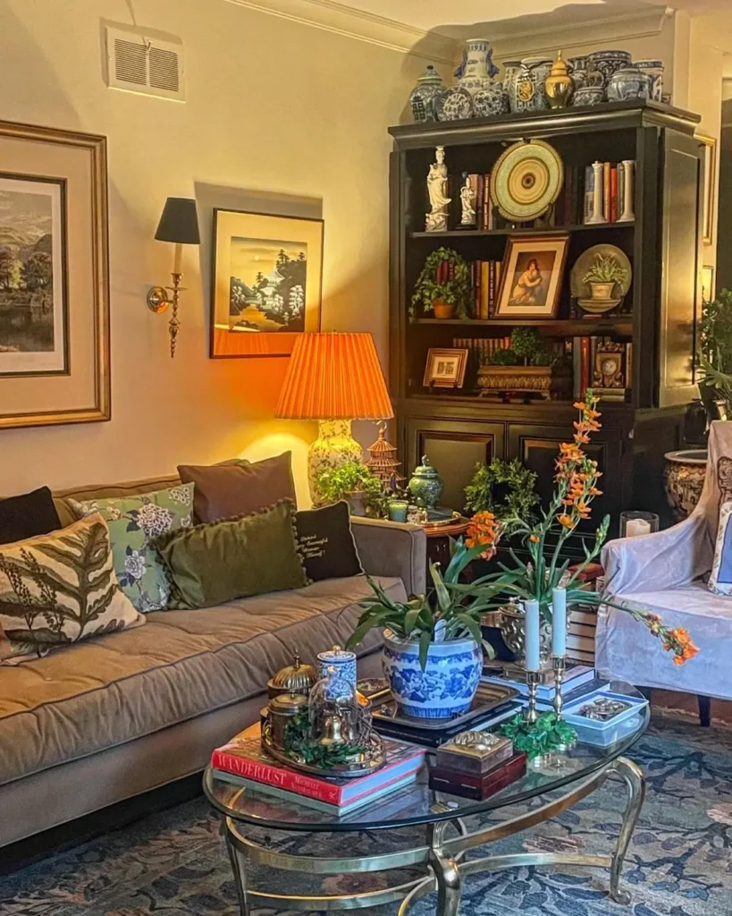 Cozy living room featuring a beige sofa adorned with colorful cushions, a lamp, and a coffee table showcasing books and plants. The bookshelf packed with decor items and framed art adds a touch of maximalist decor, creating your ultimate guide to bold living spaces.