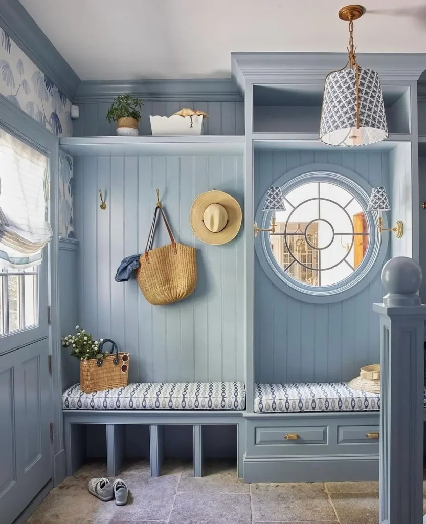 A tidy blue mudroom with built-in benches, hooks holding hats and a straw bag, a round window, patterned cushions, and decorative items on upper shelves—discover ideas in Your Ultimate Functional and Stylish Mudroom Guide.