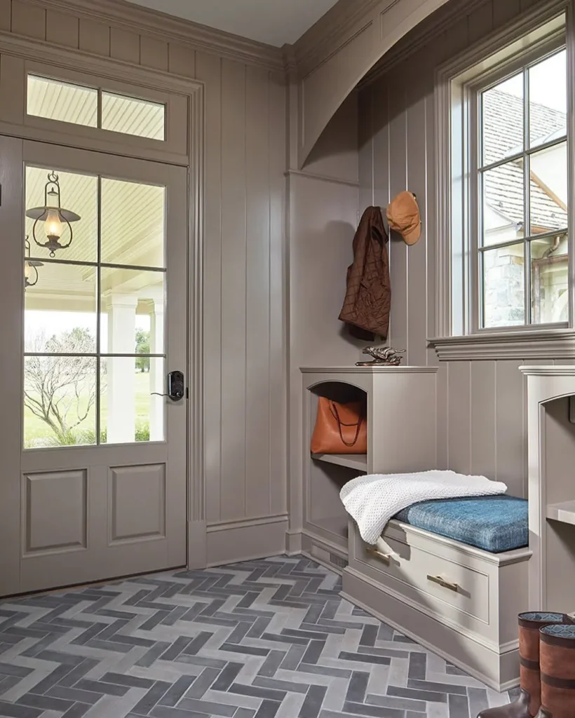 A tidy mudroom with taupe paneled walls, a bench with a blue cushion, storage drawers, and hooks for jackets and hats—discover inspiration in Your Ultimate Functional and Stylish Mudroom Guide. Natural light pours in through the glass door and window.