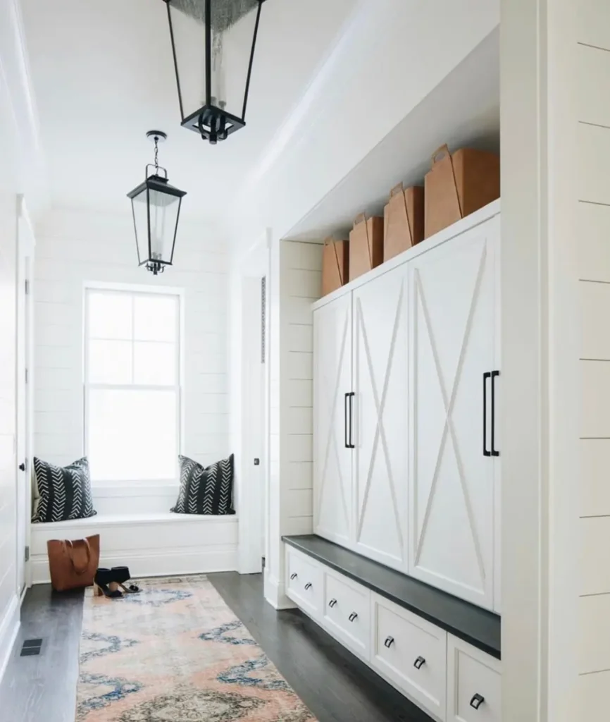 A bright hallway with white paneled walls, built-in cabinets, black handles, a window bench with pillows, brown paper bags on top, and a patterned rug on the dark floor—just like in Your Ultimate Functional and Stylish Mudroom Guide.