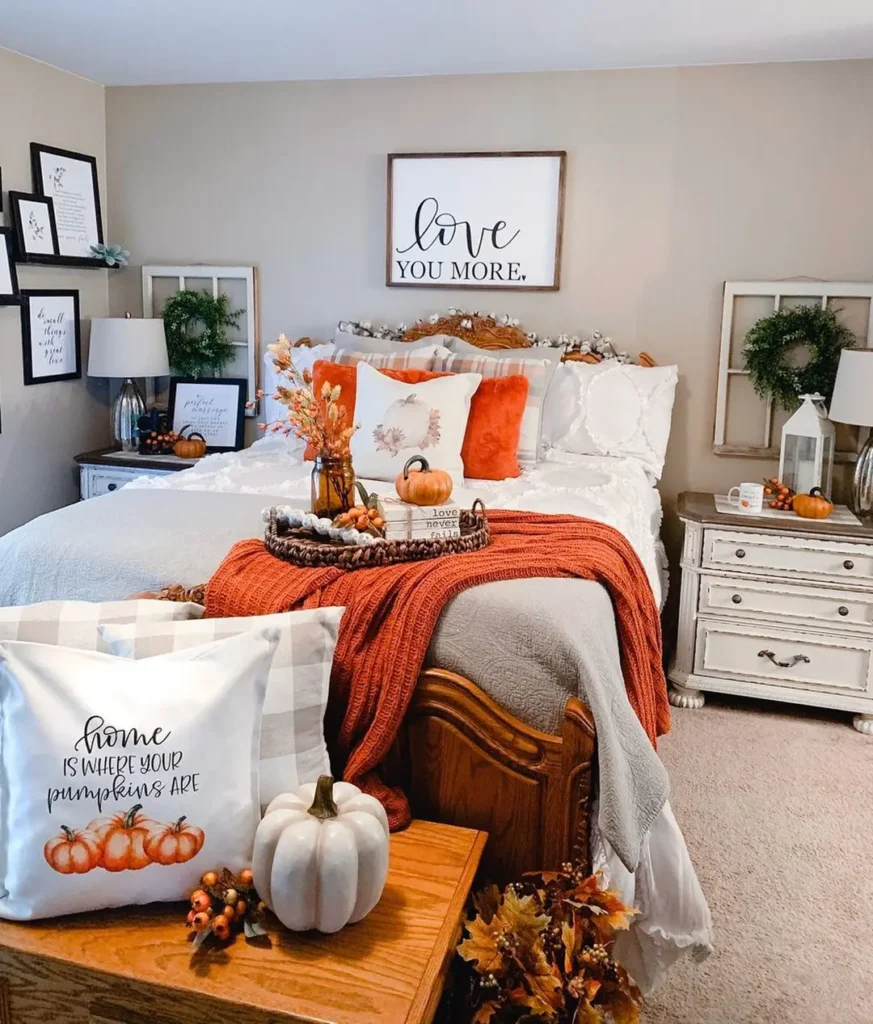A neatly decorated bedroom shows how to make your bedroom romantic this fall, featuring autumn-themed decor with orange accents, pumpkins, and cozy blankets on a white bed with framed art and pillows.