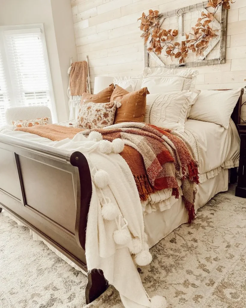 A neatly made bed with layered pillows and blankets in earthy tones, positioned against a light wood accent wall with a decorative leaf wreath above the headboard, is the perfect inspiration for How to Make Your Bedroom Romantic This Fall.