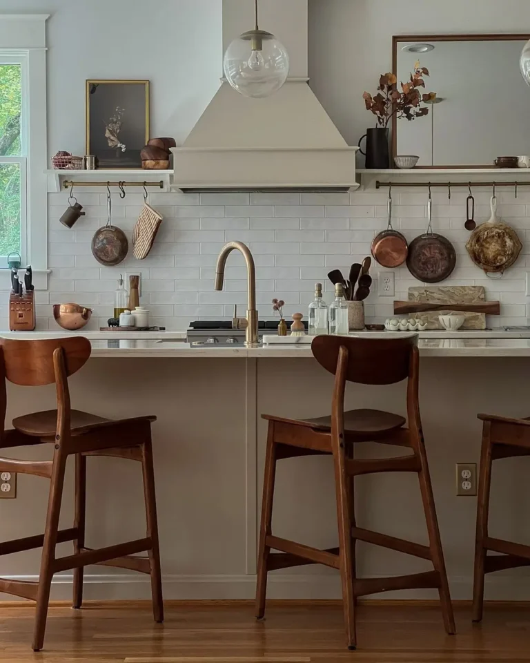 A modern kitchen with wooden stools, a white countertop, open shelves, and hanging copper pans—perfect inspiration for the 10 Best Holiday Kitchen Gifts for the Home Cook, with utensils neatly arranged against a white subway tile backsplash.