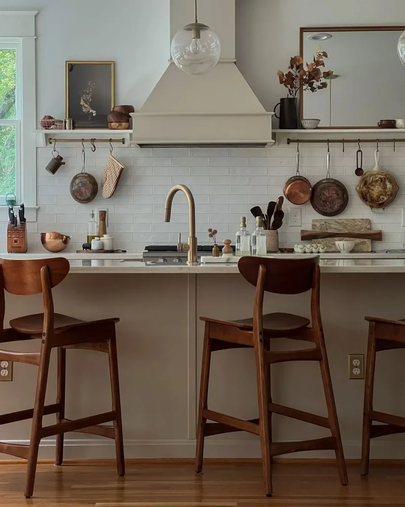 A modern kitchen with wooden stools, a white countertop, open shelves, and hanging copper pans—perfect inspiration for the 10 Best Holiday Kitchen Gifts for the Home Cook, with utensils neatly arranged against a white subway tile backsplash.