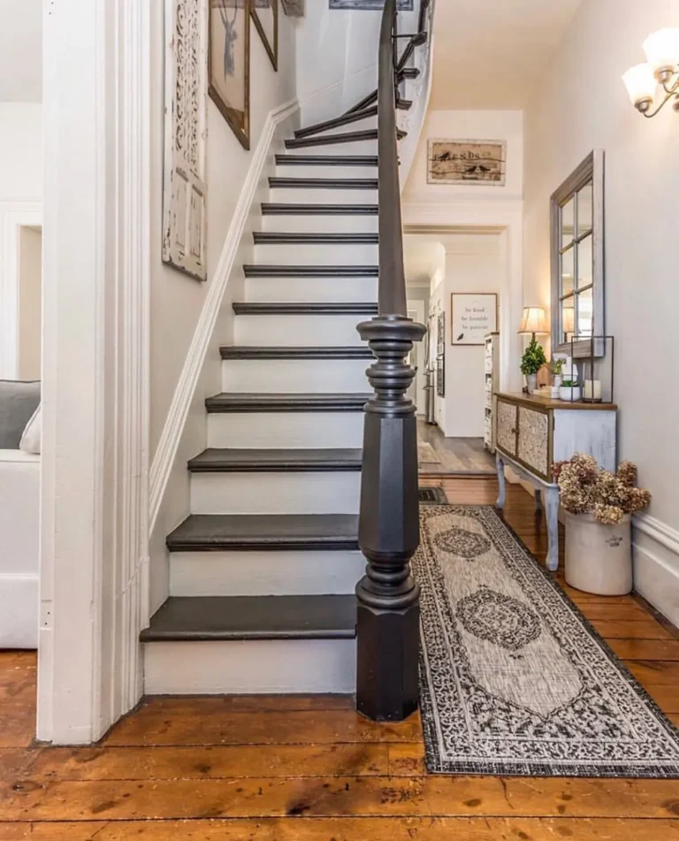 A wooden staircase with black handrail, ornate newel post, and framed art leads up from a hallway featuring a patterned rug, console table, decorative accents, and small entryway furniture chosen to fit perfectly.