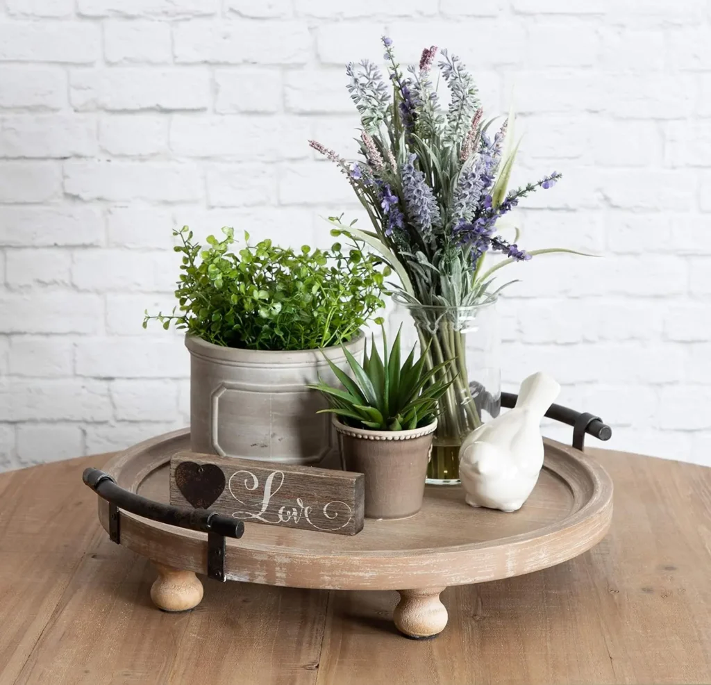 A round wooden tray holds a potted plant, a glass vase with flowers, a small potted succulent, a white ceramic bird, and a "Love" sign—a perfect touch of rustic coffee table decor on the wooden table.