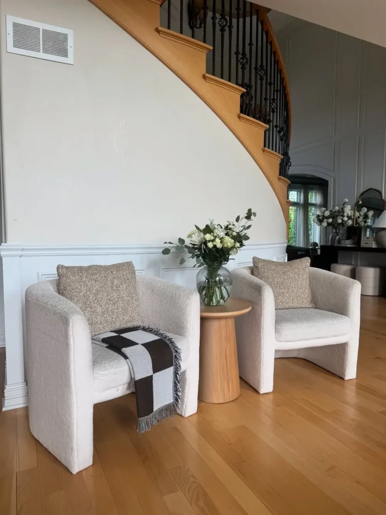 Two upholstered armchairs with beige cushions sit beside a small wooden table holding a vase of white flowers, creating an organic modern look in front of a staircase with a wooden banister and natural elements throughout the space.